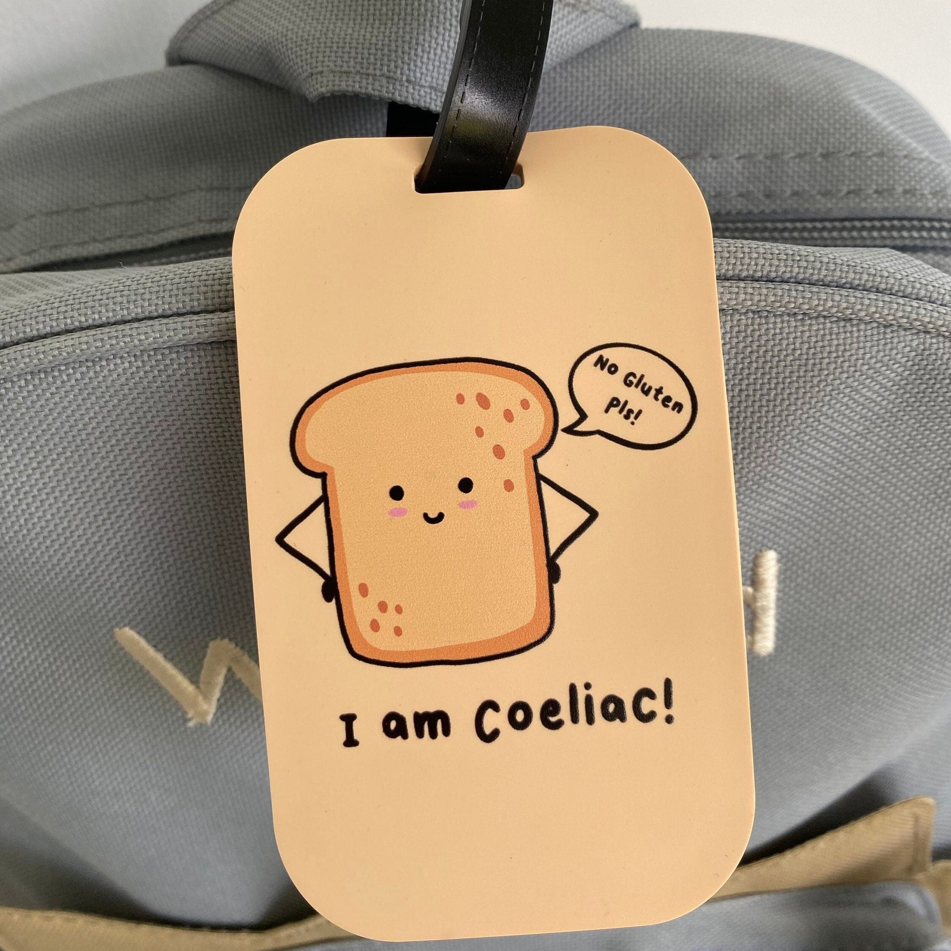 A beige luggage tag with a cartoon slice of toast and the text 'I am Coeliac!' hanging from a black strap on a gray bag.