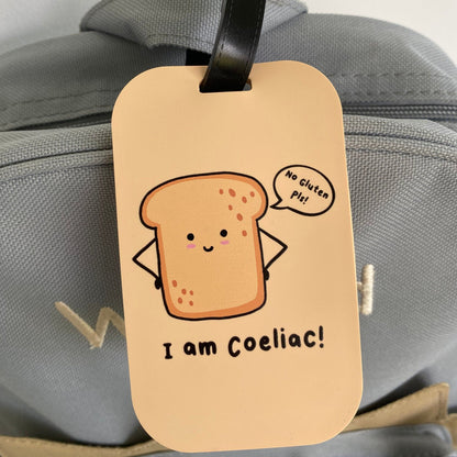 A beige luggage tag with a cartoon slice of toast and the text 'I am Coeliac!' hanging from a black strap on a gray bag.