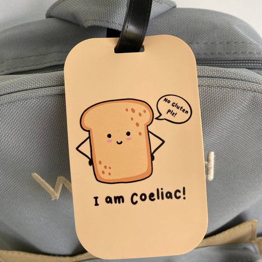 A beige luggage tag with a cartoon slice of toast and the text 'I am Coeliac!' hanging from a black strap on a gray bag.