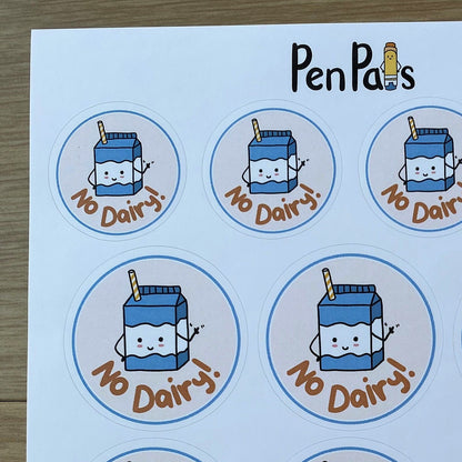 A sheet of round stickers featuring a cute cartoon milk carton with the text 'No Dairy!' in brown, arranged on a white sheet over a wood surface. The brand name 'PenPals' is printed at the top.