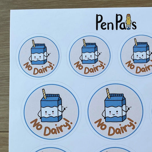 A sheet of round stickers featuring a cute cartoon milk carton with the text 'No Dairy!' in brown, arranged on a white sheet over a wood surface. The brand name 'PenPals' is printed at the top.