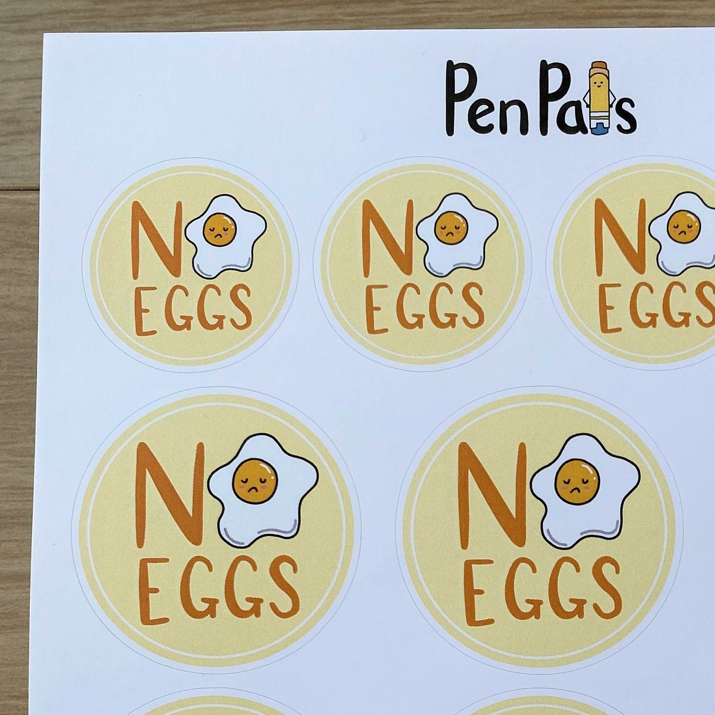 A sheet of round stickers printed with the phrase 'NO EGGS' and a cartoon fried egg, branded 'PenPal's' at the top, shown on a light wood surface.