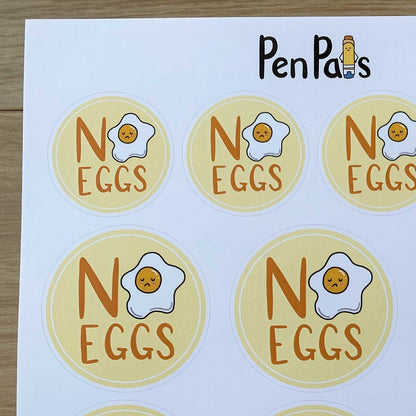 A sheet of round stickers printed with the phrase 'NO EGGS' and a cartoon fried egg, branded 'PenPal's' at the top, shown on a light wood surface.