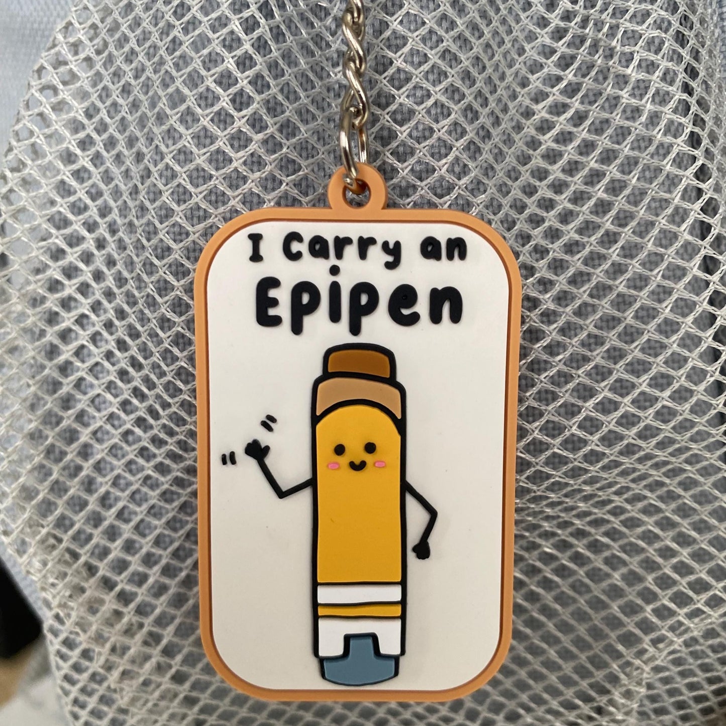 A rectangular enamel keychain charm with rounded corners featuring a cartoon yellow pencil character and the text 'I Carry an Eipepen' on a white background with an orange border, hanging from a silver chain against a gray mesh surface.