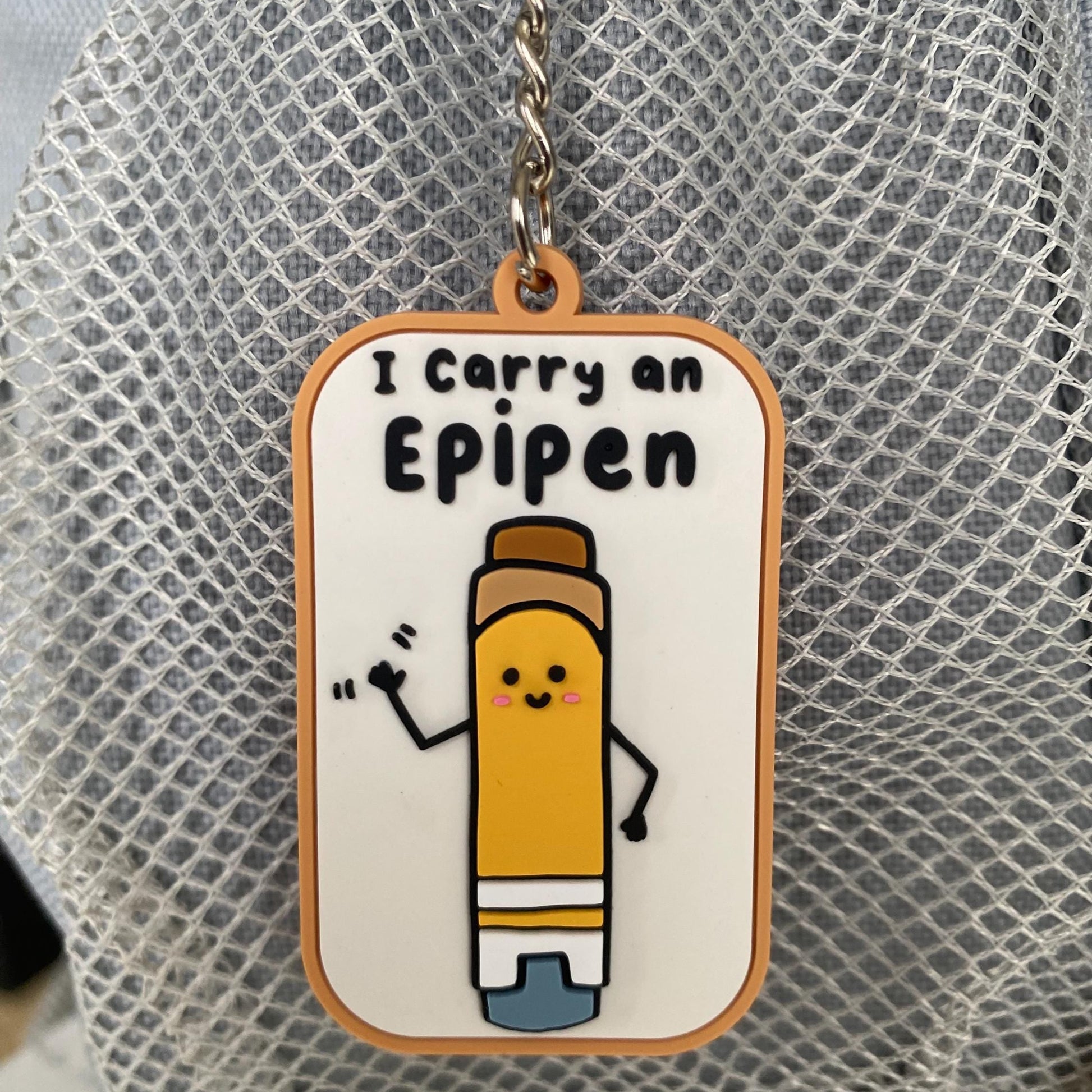 A rectangular enamel keychain charm with rounded corners featuring a cartoon yellow pencil character and the text 'I Carry an Eipepen' on a white background with an orange border, hanging from a silver chain against a gray mesh surface.