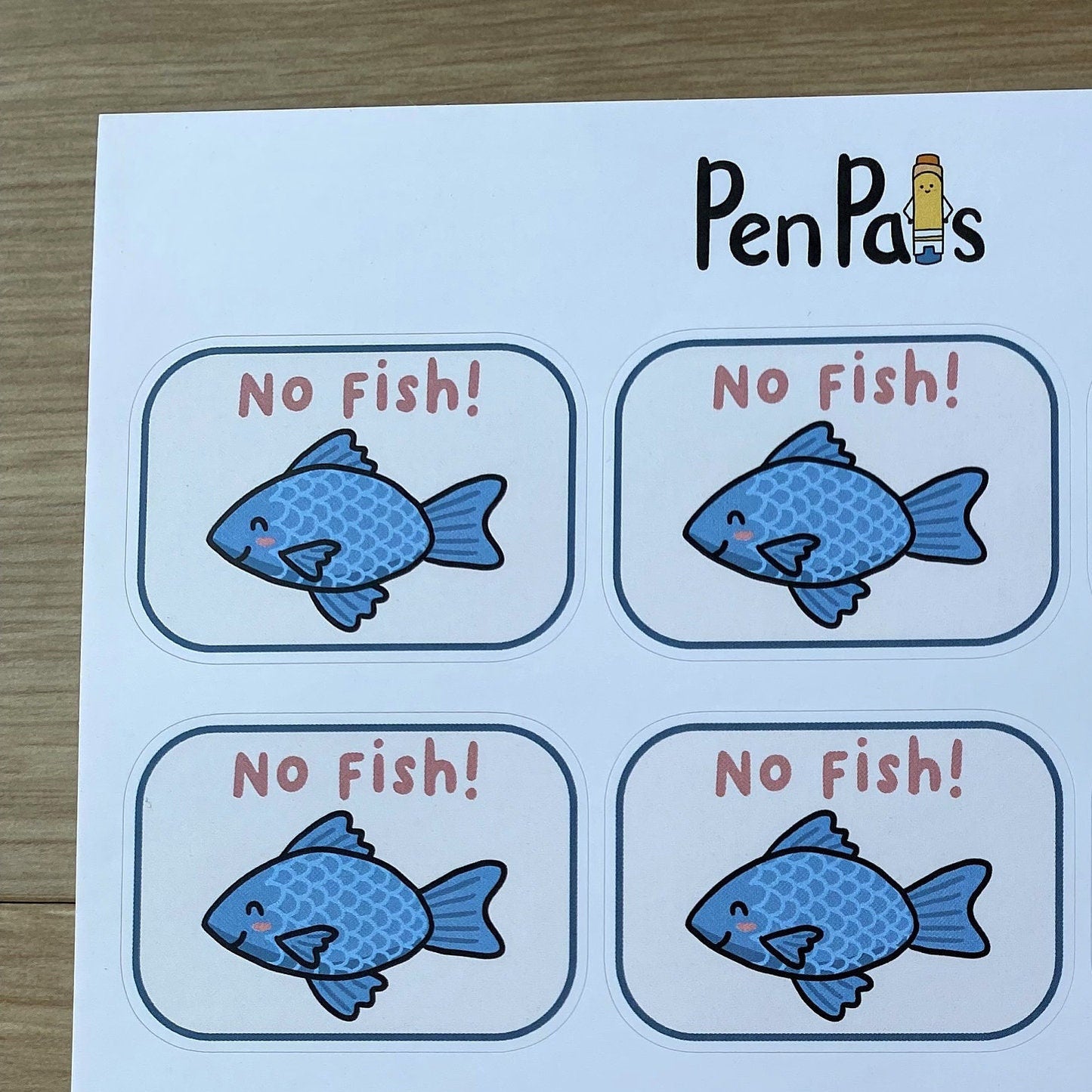 A sheet of printed labels or stickers with the brand name PenPals at the top, featuring nine cartoon blue fish illustrations and the text 'No Fish!' in red inside rounded white boxes, placed on a light wood surface.
