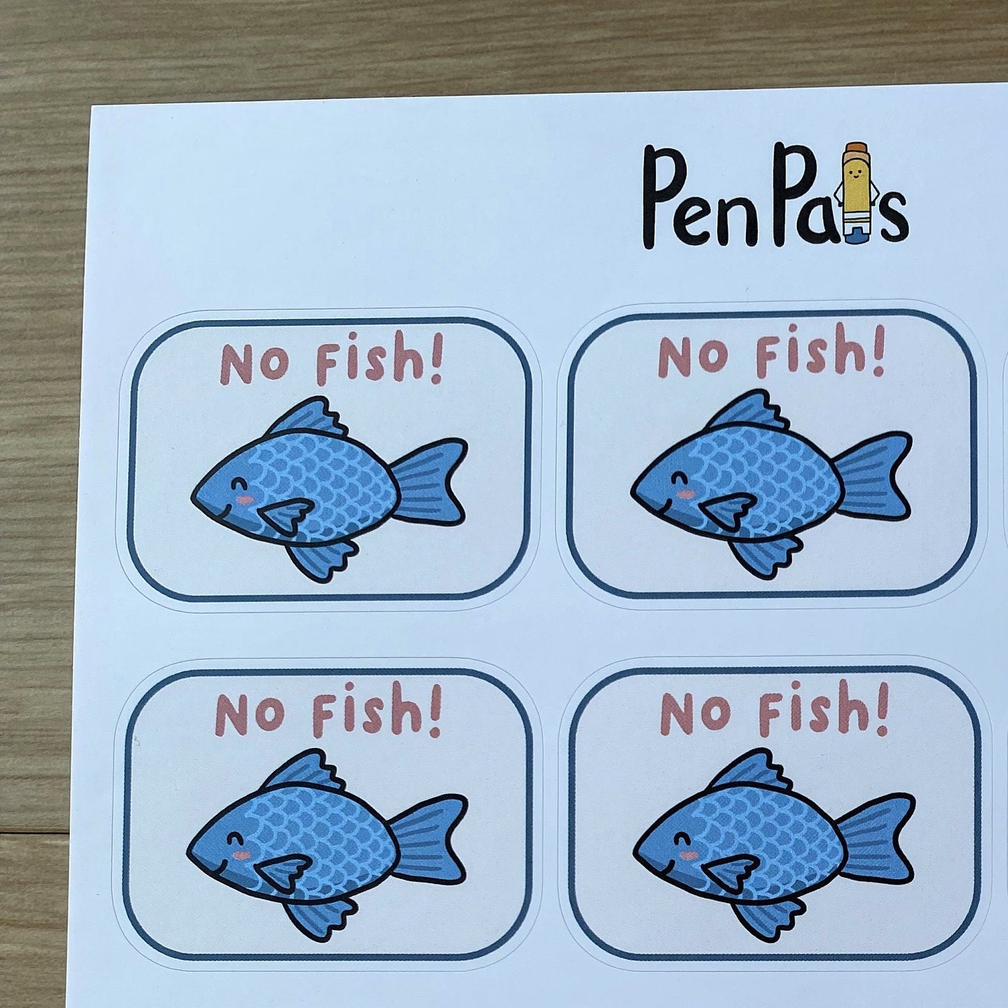 A sheet of printed labels or stickers with the brand name PenPals at the top, featuring nine cartoon blue fish illustrations and the text 'No Fish!' in red inside rounded white boxes, placed on a light wood surface.