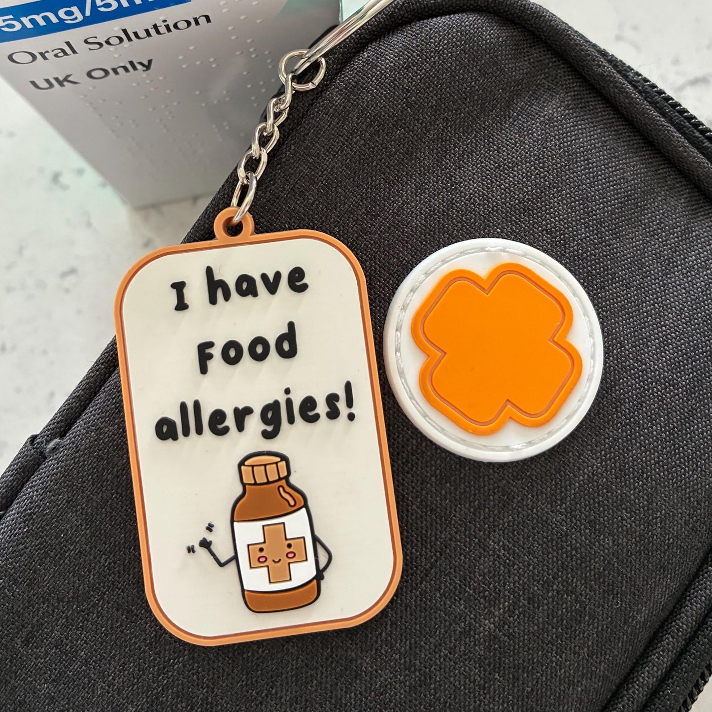 A close-up of a dark gray zippered pouch with a keychain tag that reads 'I have Food allergies!' and shows a small orange clover-shaped charm, with a Cetirizine Hydrochloride box partially visible in the background.