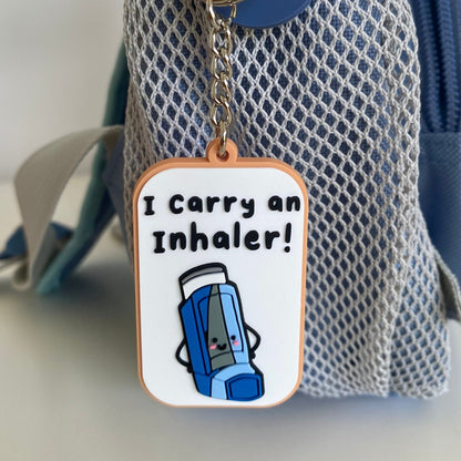 A close-up of a blue and gray mesh bag with a hanging keychain tag that reads 'I Carry an Inhaler!' with a cartoon blue inhaler illustration.