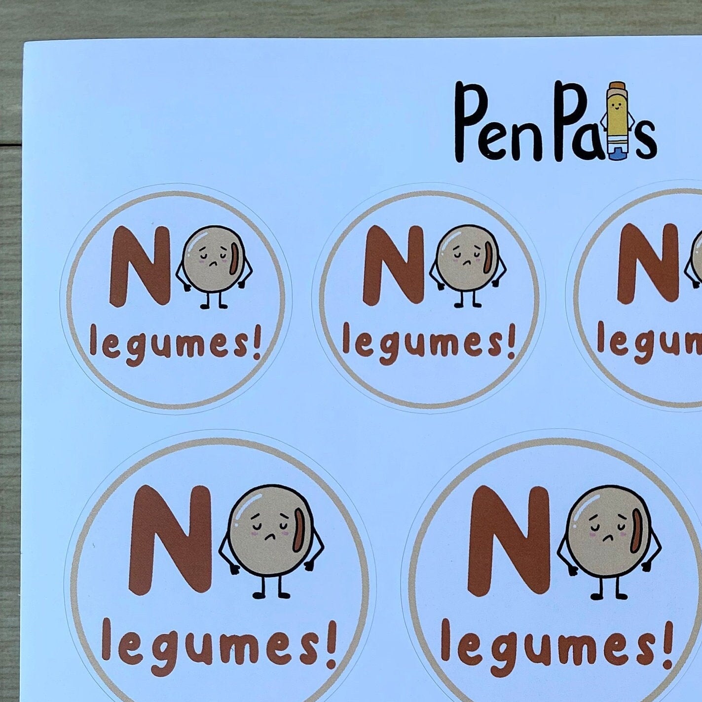 A sheet of circular labels printed with the text 'NO legumes!' and a cartoon potato character, shown on a light wood surface. The brand name 'PenPals' appears at the top.