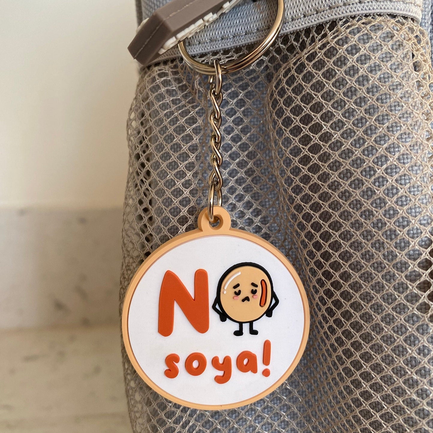 A circular keychain charm with a cartoon face and the text 'No soya!' hanging from a metal ring on a gray mesh bag.