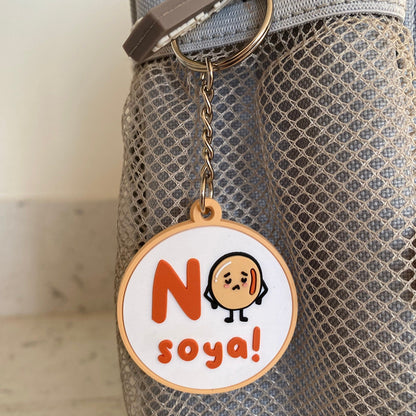A circular keychain charm with a cartoon face and the text 'No soya!' hanging from a metal ring on a gray mesh bag.