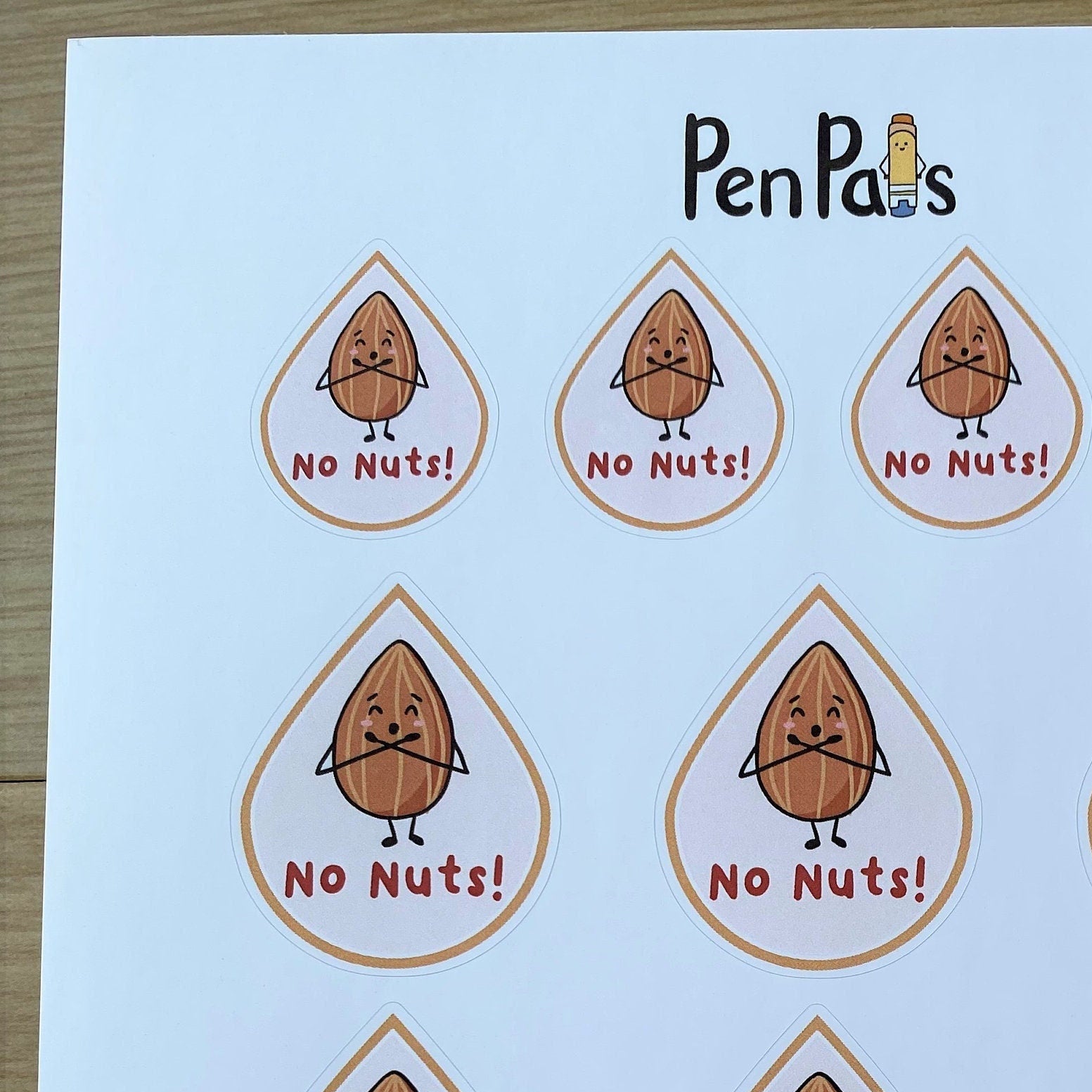 A white printed sheet featuring twelve cartoon nut illustrations inside teardrop-shaped outlines, each with the text 'No Nuts!' and the brand name 'Pen Pals' at the top, photographed on a wood surface.