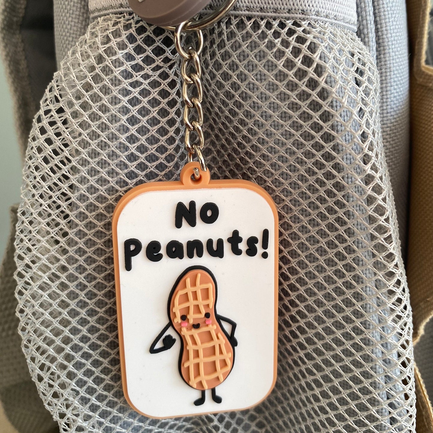 A close-up of a gray mesh bag with a hanging keychain tag that reads 'No Peanuts!' and shows a cartoon peanut character. 
