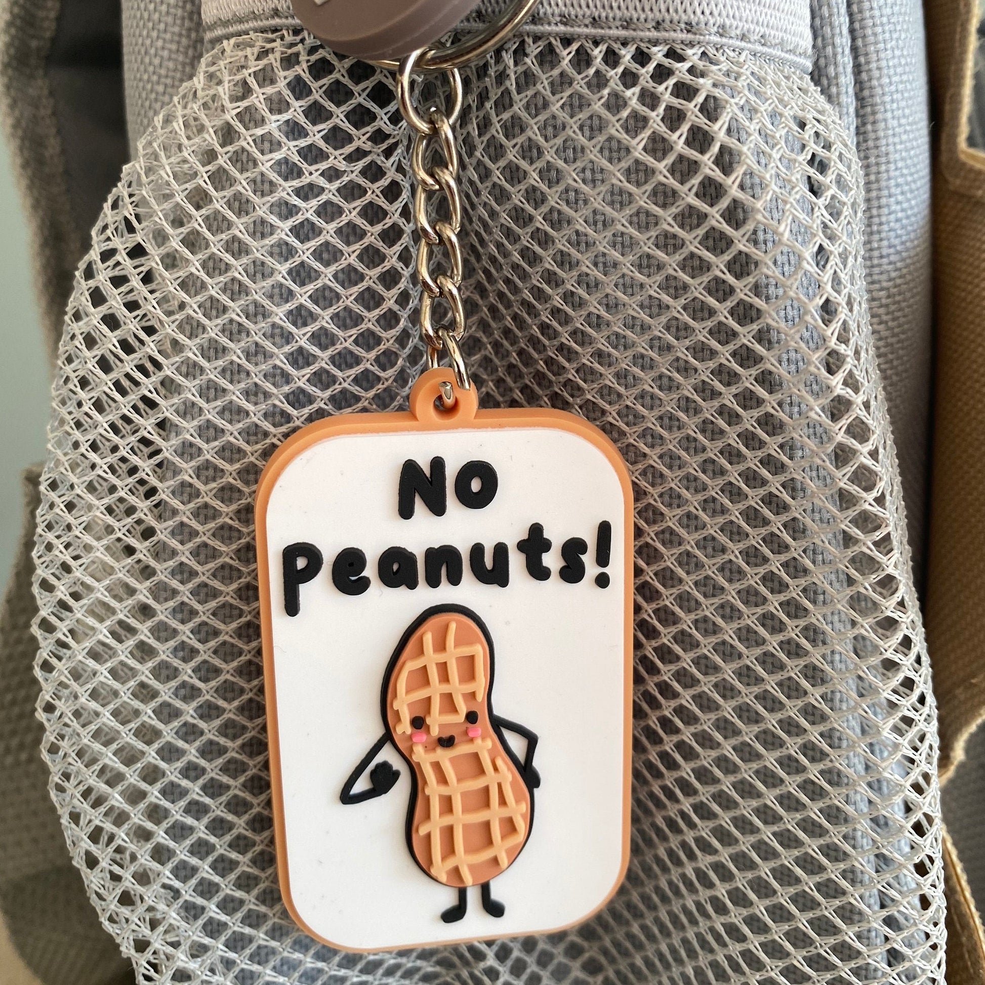 A close-up of a gray mesh bag with a hanging keychain tag that reads 'No Peanuts!' and shows a cartoon peanut character. 