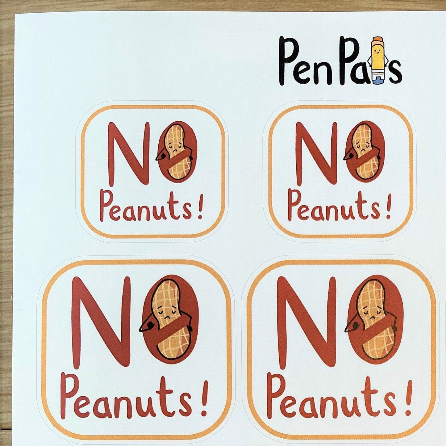 A sheet of 12 small square stickers with the text 'NO Peanuts!' and a peanut icon, printed on a white card with the brand name PenPals at the top, photographed on a wood surface.