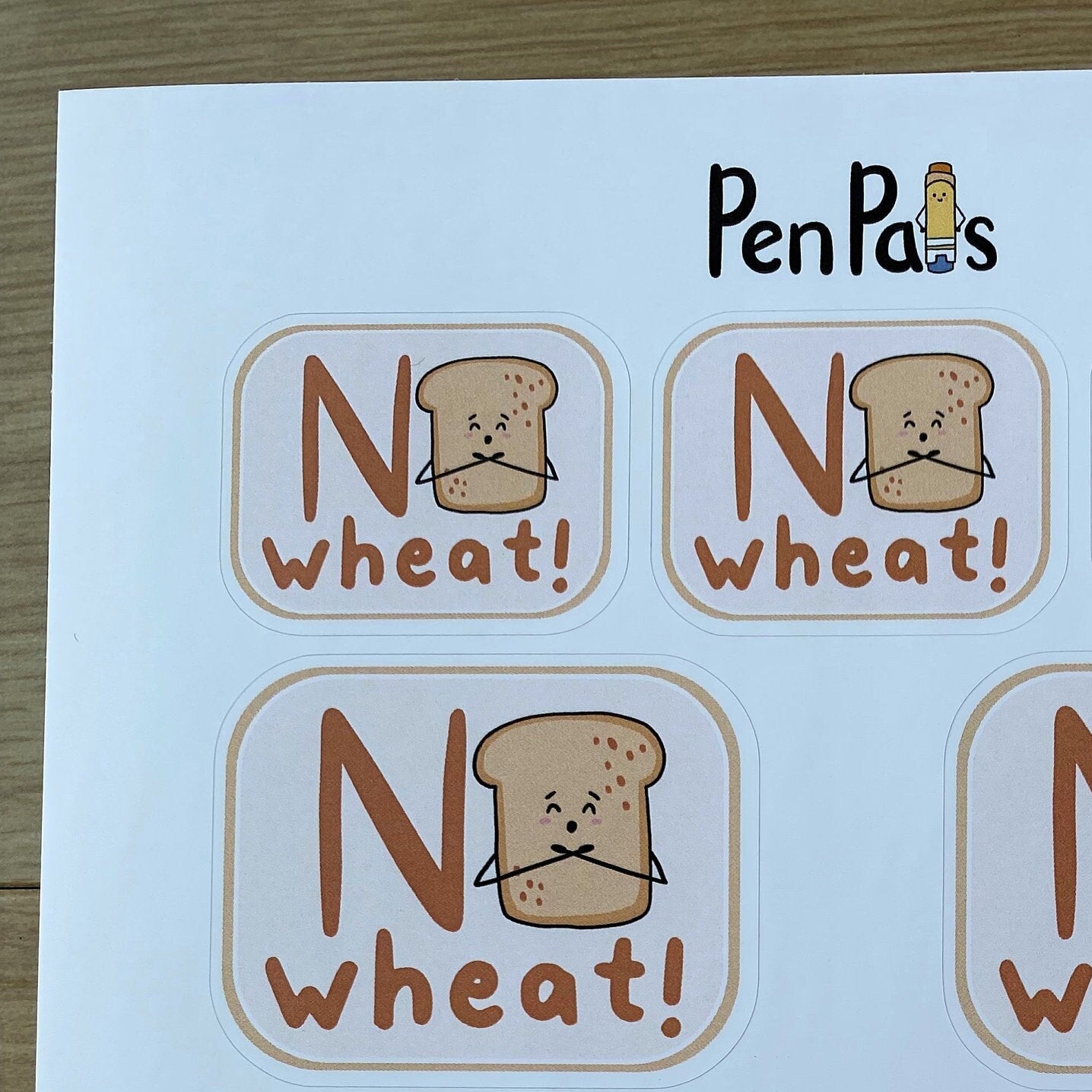 A sheet of printed labels or stickers featuring the phrase 'No wheat!' with a cartoon slice of bread and the brand name 'PenPal's' at the top, photographed on a wood surface.