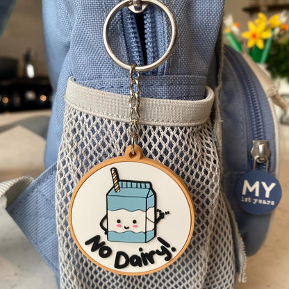 A close-up of a light blue fabric bag with a front mesh pocket and a round hanging charm that reads “No Dairy!” with a cartoon milk carton. A small blue tag on the bag reads “MY 25 YEARS.