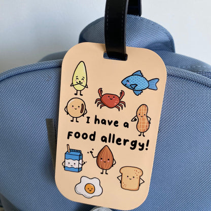 A beige luggage tag with a black strap hanging on a blue backpack. The tag features a cartoon-style food allergy message reading 'I have a food allergy!' surrounded by cute illustrated food items like a banana, crab, fish, potato, peanut, milk carton, almond, egg, and toast.