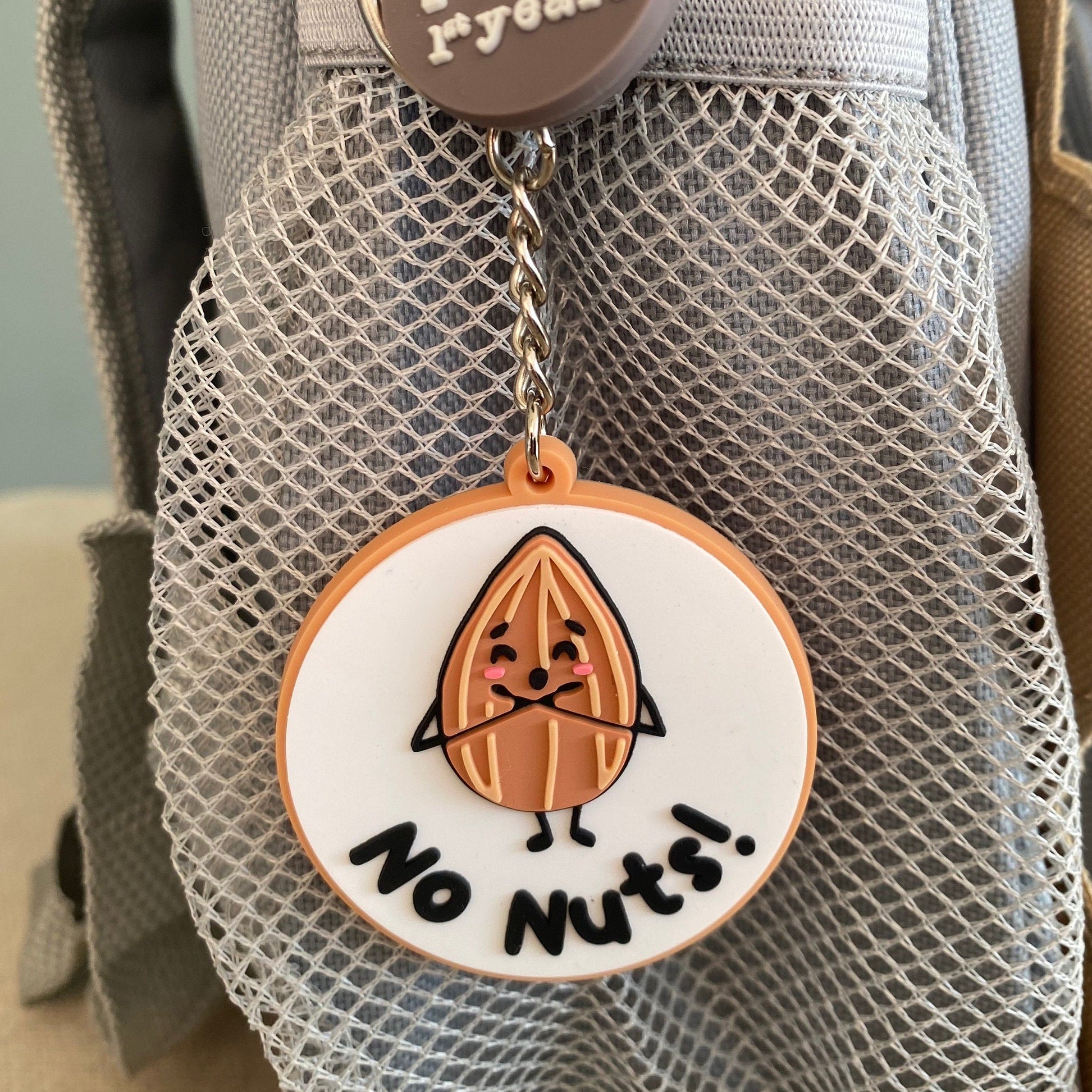 A close-up of a gray mesh backpack pocket with a round metal keychain charm. The charm has a white face with a cartoon nut character and the text 'No Nuts!' in black,