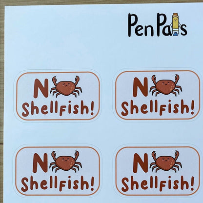 A sheet of round stickers printed with the phrase 'No Shellfish!' in brown lettering, featuring a small orange crab illustration between the words. The brand name 'PenPals' appears at the top of the sheet.