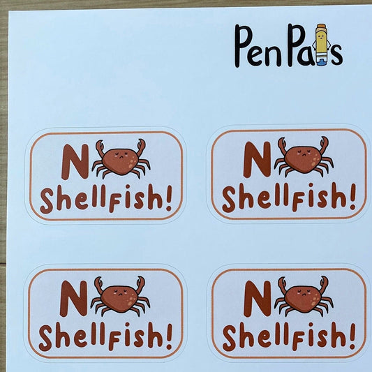 A sheet of round stickers printed with the phrase 'No Shellfish!' in brown lettering, featuring a small orange crab illustration between the words. The brand name 'PenPals' appears at the top of the sheet.