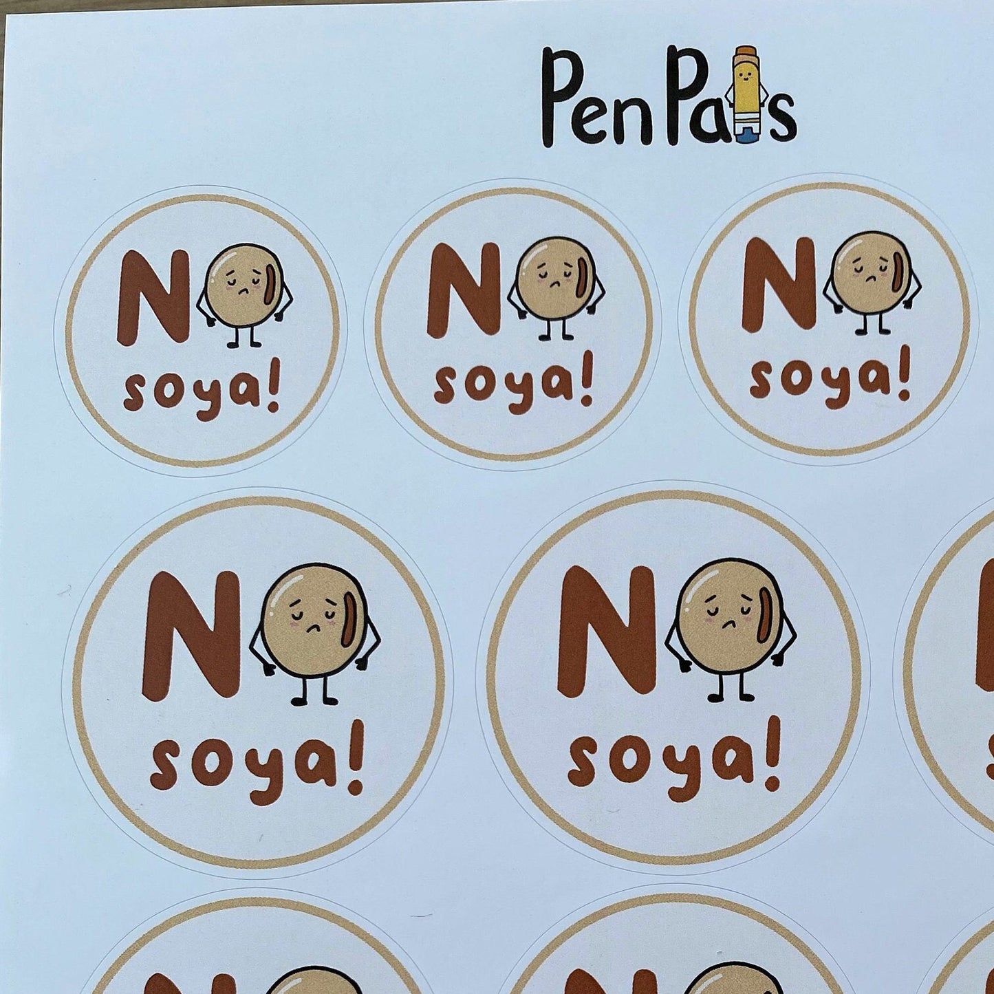 A sheet of circular printed labels or stickers with the text 'No soya!' and a cartoon potato character, placed on a wooden surface. The brand name 'PenPas' is printed at the top.